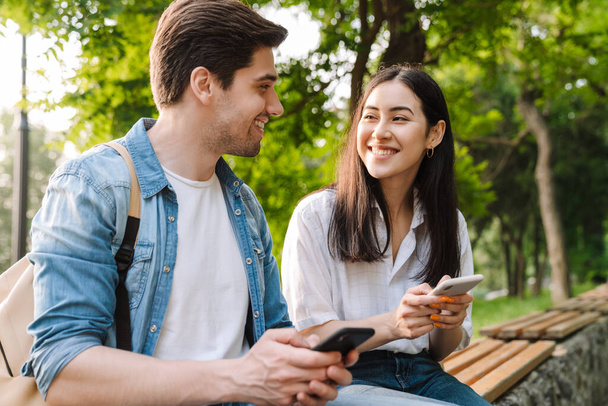 Image of pleased multicultural couple using mobile phones and smiling while sitting on bench in park - Fotografie, Obrázek