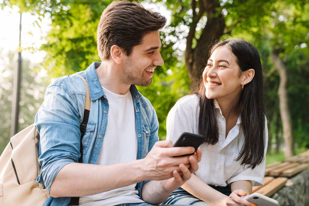 Image of pleased multicultural couple using mobile phones and smiling while sitting on bench in park - Photo, image
