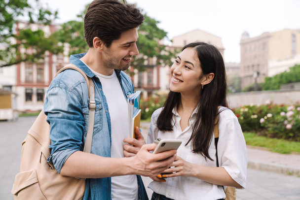 Image of joyful multicultural student couple talking and using cellphone while walking on boulevard - Valokuva, kuva