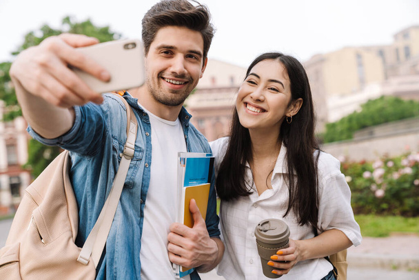 Image of joyful multicultural student couple taking selfie photo on cellphone while walking on boulevard - Photo, Image