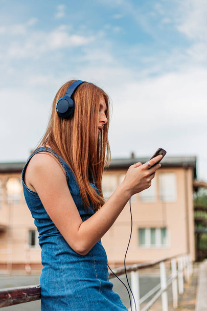 Cute, ginger girl streaming music on her phone. Headphones on her head, school yard - 写真・画像