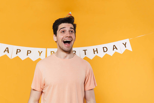 Photo of excited young man in party cone expressing surprise on camera isolated over yellow background - Photo, image