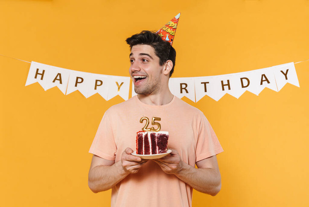 Photo of excited caucasian man in party cone holding birthday cake and smiling isolated over yellow background - 写真・画像