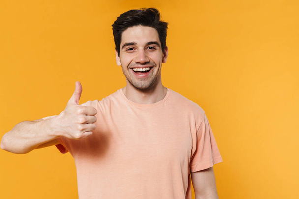 Foto van vrolijke knappe man met duim omhoog en glimlachend geïsoleerd over gele achtergrond - Foto, afbeelding