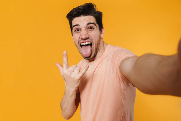 Young brunette happy man showing tongue and rock gesture isolated over yellow - Fotografie, Obrázek