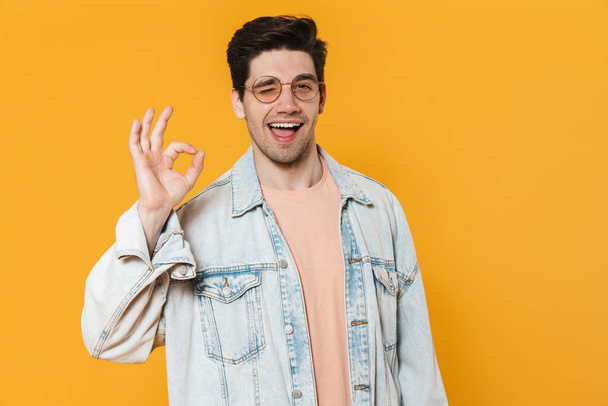 Photo of joyful young man in eyeglasses gesturing ok sign and winking isolated over yellow background - Фото, зображення
