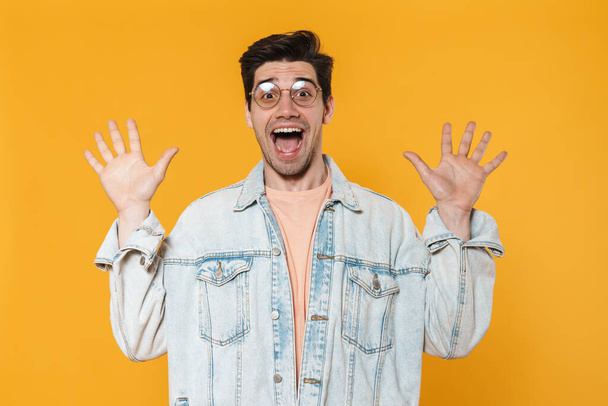 Photo of excited young man in eyeglasses expressing surprise at camera isolated over yellow background - Fotoğraf, Görsel