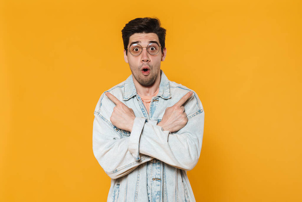 Photo of surprised young man in eyeglasses pointing fingers aside isolated over yellow background - Foto, imagen