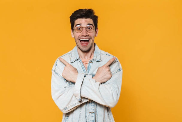 Photo of delighted young man in eyeglasses pointing fingers aside isolated over yellow background - Fotoğraf, Görsel