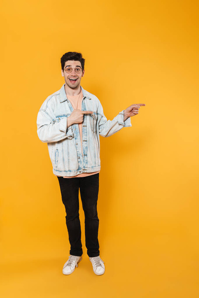 Photo of joyful young man in eyeglasses pointing fingers aside and smiling isolated over yellow background - Photo, Image