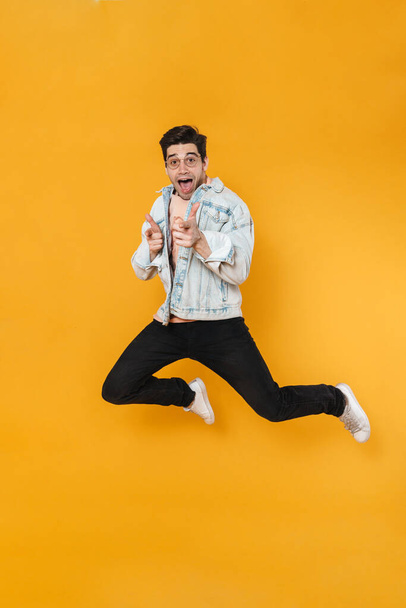 Photo of delighted young man jumping and pointing fingers at camera isolated over yellow background - Photo, image