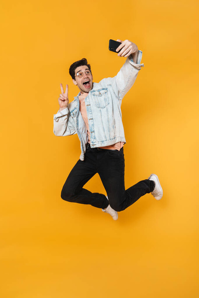 Photo of delighted man gesturing peace sign while taking selfie on cellphone isolated over yellow background - Foto, Bild