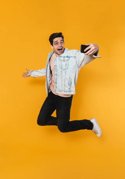 Photo of delighted caucasian man jumping while taking selfie on cellphone isolated over yellow background - Foto, afbeelding
