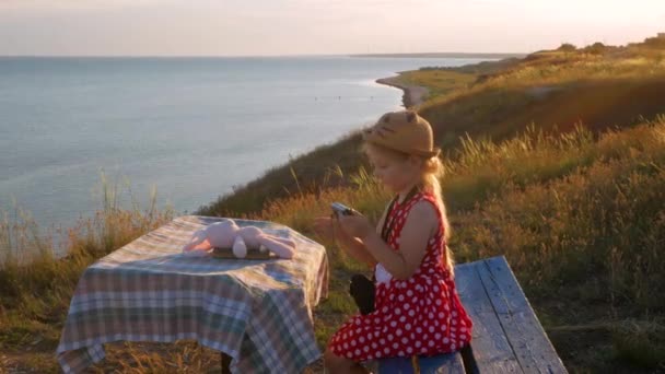 Child girl in a straw hat and dress sitting on vintage bench and taking a picture. Cute kid with soft pink rabbit toy looking at notebook in hands on sea nature lanscape background. Friendship concept - Footage, Video