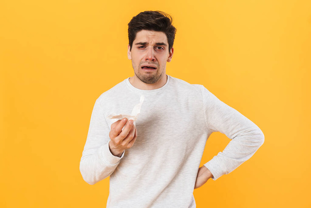 Photo of sick unhappy man with runny nose posing with tissue on camera isolated over yellow background - Фото, изображение