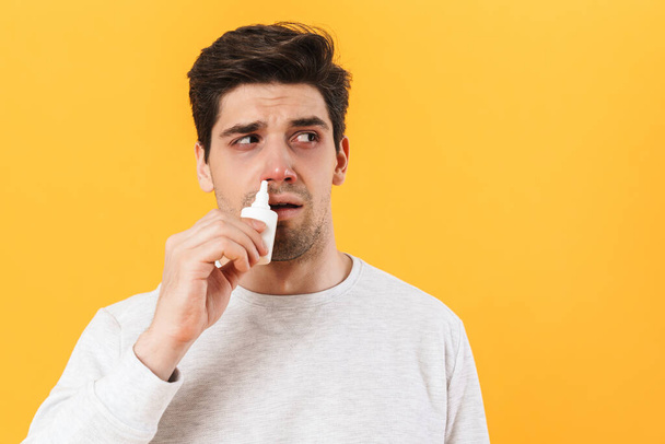 Photo of handsome unhappy man with allergy using nose drops isolated over yellow background - Photo, image