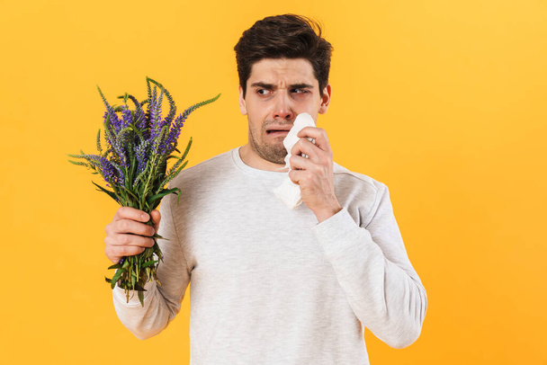 Foto de hombre guapo infeliz con alergia posando con flores aisladas sobre fondo amarillo - Foto, Imagen