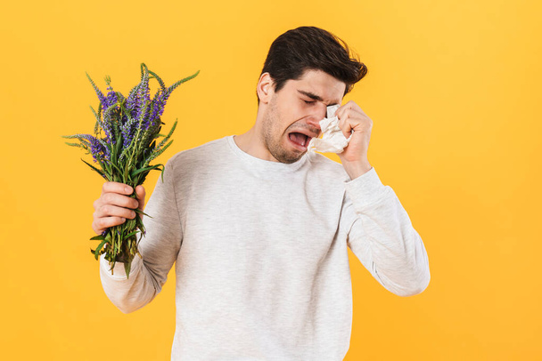 Photo of handsome unhappy man with allergy posing with flowers isolated over yellow background - 写真・画像