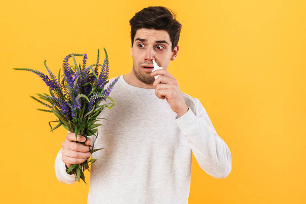 Photo of unhappy man with allergy using nose drops while holding flowers isolated over yellow background - Фото, изображение