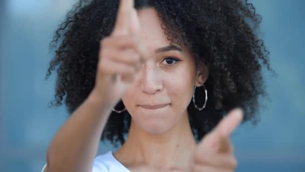 Charming stylish African American teen girl with afro hairstyle makes hand pistols hit goal, pretends to shoot gun from fingers smile happily toothy. Beautiful young woman flirts amusingly confidently - Materiaali, video