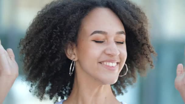 Happy excited African American girl shakes her head with curly hair, smiles happily and raises her fists in victory gesture. A young woman is delighted with the good news, sincere emotions, success. - Felvétel, videó