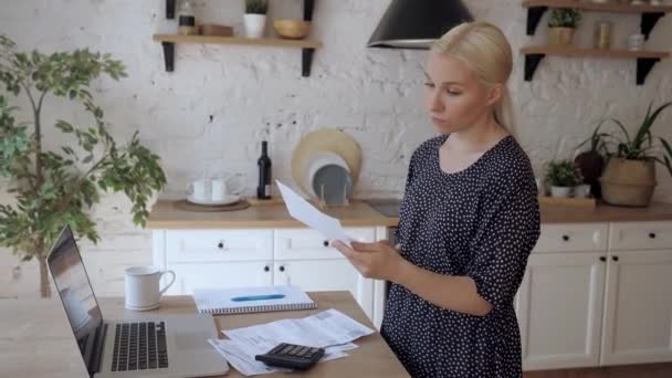 A young woman standing in the kitchen opens the utility bill and is very upset - Materiał filmowy, wideo