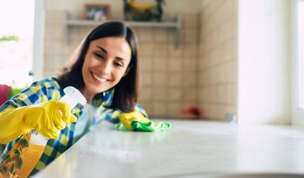 Happy beautiful young woman in yellow gloves is cleaning the kitchen with special equipment and spray. Housework and cleaning stuff - Foto, Bild