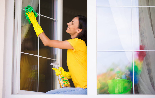 Beautiful young happy woman is using a duster and a spray while cleaning a window in the house. Homework concept - Foto, Imagen
