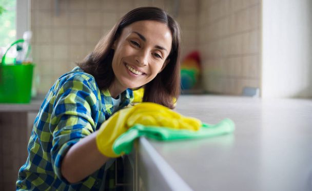 Happy beautiful young woman in yellow gloves is cleaning the kitchen with special equipment and spray. Housework and cleaning stuff - Foto, imagen