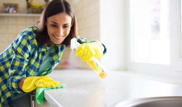 Happy beautiful young woman in yellow gloves is cleaning the kitchen with special equipment and spray. Housework and cleaning stuff - Photo, image
