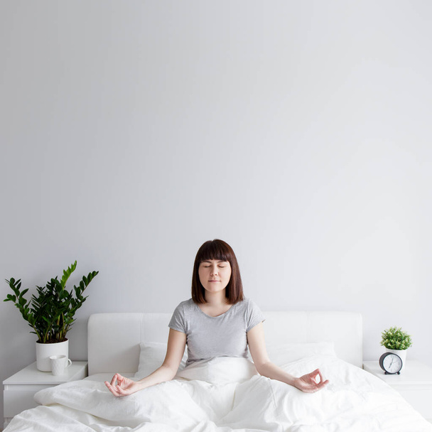 happy woman sitting on bed in yoga pose at home, copy space over white wall background - Фото, изображение