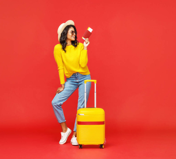 Teljes test trendi etnikai nő útlevéllel és jegyek mosolygós kamera poggyász a nyári vakáció alatt ellen piros backdro - Fotó, kép