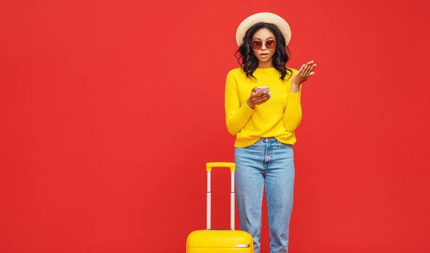 Sorprendido turista étnico ver horarios de vuelo en su teléfono inteligente y soñando con vacaciones mientras abraza maleta contra backdro rojo - Foto, imagen
