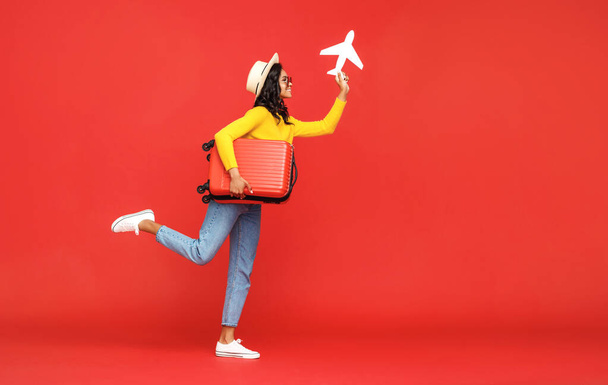 Cuerpo completo étnico femenino en sombrero y gafas de sol saltando con la maleta y jugando con el avión de juguete contra el backdro rojo - Foto, imagen