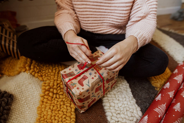 Woman wrapping red christmas gift box under christmas tree  in modern room, close up. Happy girl in sweater sitting and preparing christmas presents. Happy holidays - Фото, зображення