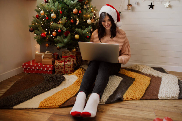 現代的な部屋の照明や装飾品とクリスマスツリーの下にラップトップで座ってスタイリッシュな若い女性。サンタの帽子の幸せな女の子はノートパソコンで作業し、クリスマスの販売で買い物. - 写真・画像