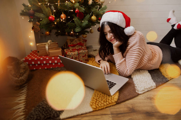 Elegante joven sentada con portátil en la alfombra bajo el árbol de Navidad con regalos en la habitación moderna. Chica feliz en sombrero de santa trabajando o de compras en las ventas de Navidad. Felices fiestas - Foto, Imagen