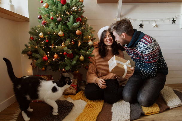 Mutlu noeller. Noel ağacının altında ışıklarla Noel hediyelerini değiş tokuş eden mutlu bir çift. Genç bir aile sürpriz hediyeler sunuyor, neşeli modern odada mutlu anlar.. - Fotoğraf, Görsel