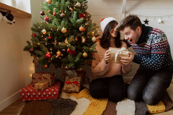 Feliz pareja emocionada intercambiando regalos de Navidad bajo el árbol de Navidad con luces. Joven familia sosteniendo regalo sorpresa, feliz momento en la habitación moderna festiva. Feliz Navidad. - Foto, imagen