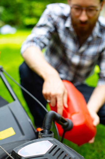 Man refilling a fuel tank in a petrol lawn mower with a red plastic canister with a funnel, close up and focus on a funnel. Gardening, greenworks, using a gasoline lawnmower in a garden on a sunny day - Foto, imagen