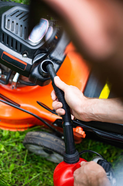 Man refilling a fuel tank in a petrol lawn mower with a red plastic canister with a funnel, close up on a hand with a funnel. Gardening, using a gasoline lawn mower in a garden on a sunny day - Foto, Imagem