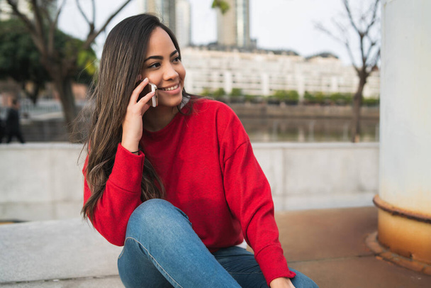 Portrait of young latin woman talking on the phone while sitting outdoors. Urban concept. - Foto, Imagem