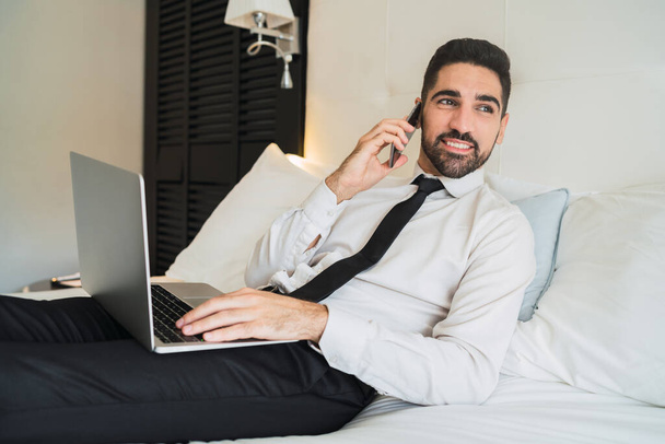 Portrait of young businessman lying on bed and talking on the phone while working on his laptop at the hotel room. Business travel concept. - Foto, imagen
