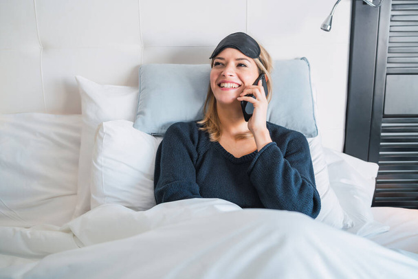 Portret van een jonge vrouw die telefoneert terwijl ze op bed ligt in de hotelkamer. Reis- en leefstijlconcept. - Foto, afbeelding
