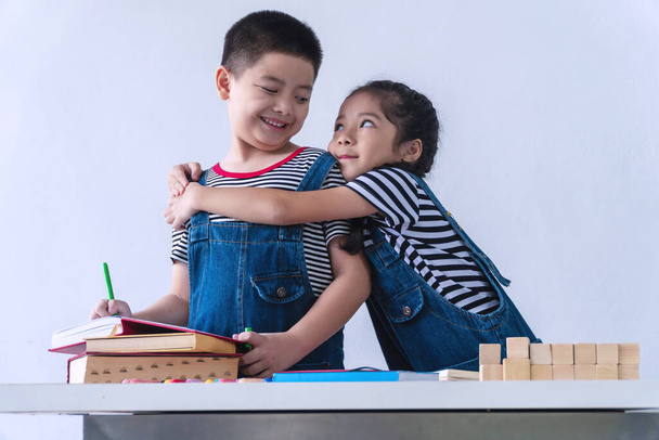 Невинность маленькая милая девочка улыбается с деревянным блоком и книги на столе, обнимая своего брата на белом фоне. Братство обнимает друг друга, чувствуя счастье. Концепция родственных отношений. - Фото, изображение