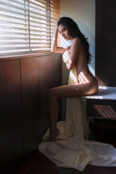Attractive sexy woman hold white thin pajamas sitting near window looking at something.Charming lonely sexy girl feels relaxation in the room. Glamour women long hair slim body gets topless or nude. - Photo, Image