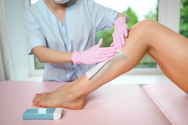 Woman with long tanned perfect legs and smooth skin having wax stripe depilation hair removal procedure on legs in beauty salon. Beautician in blue robe, pink gloves. Body care, epilation spa concept. - Foto, immagini
