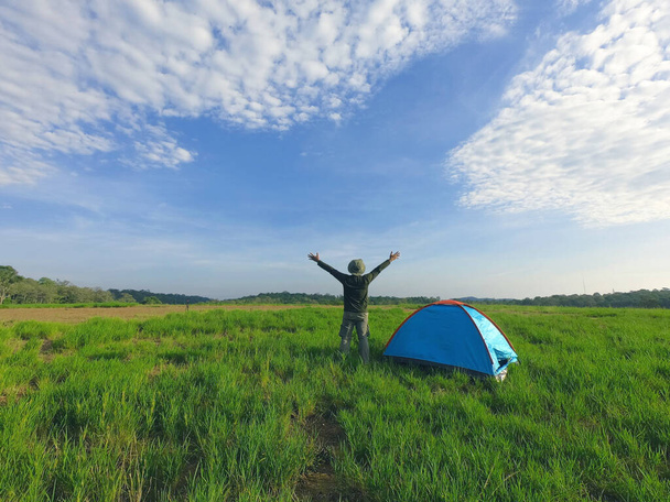 Costas de viajante homem de pé ao lado da tenda no prado, enquanto levantar a mão com atmosfera céu na frente da manhã dele. Conceito de estilo de vida aventura
. - Foto, Imagem