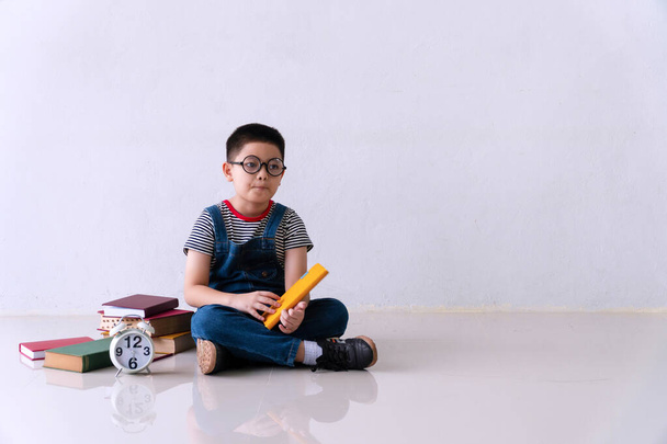 A kis cuki fiú szemüveget visel, ül a padlón egy halom könyvvel, ceruzával és órával, miközben néz valamit, aminek van másolási helye. Iskolásfiú házi feladatot rajzol otthon. Fekete-iskola koncepció. - Fotó, kép