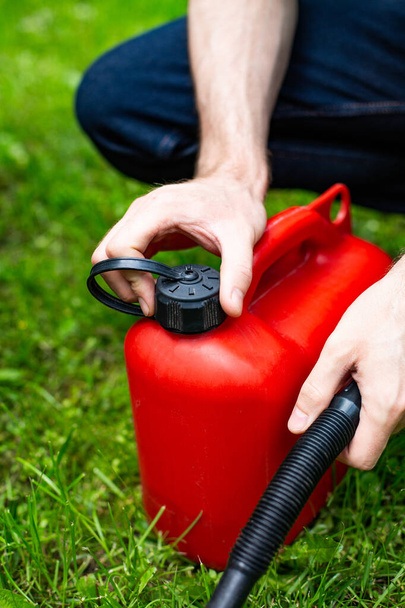Vertical view of a male hand opening a red plastic fuel canister with a funnel to refill a petrol lawn mower, close up. Gardening, greenworks, using a gasoline lawnmower in a garden on a sunny day. - Fotoğraf, Görsel